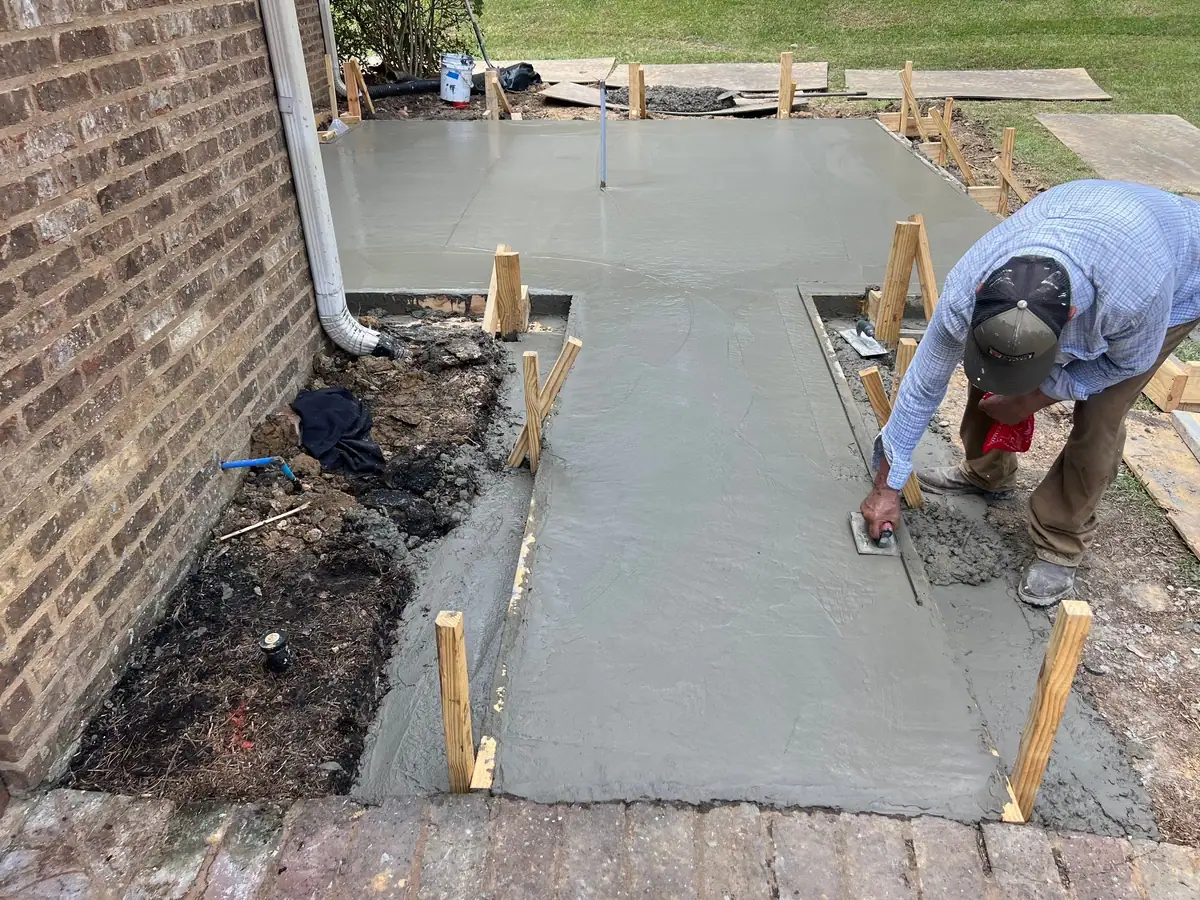 Finisher working fresh concrete on a new outdoor living patio walk beside a brick house